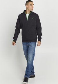 Felpa zip grigia scura con dettagli verdi e logo, abbinata a jeans azzurri e sneakers nere per un look casual.
