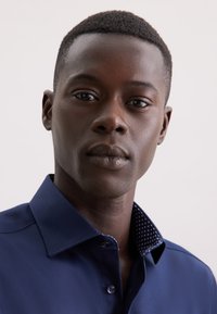 Camicia blu navy con colletto strutturato. L'interno del colletto presenta un motivo a pois. Il tessuto appare liscio e ben aderente.