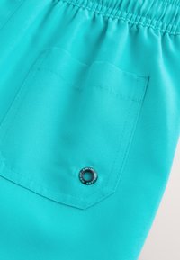 Türkiissinised ujumispüksid elastse vöökohaga, millel on tagatasku ja hõbedane logo-detailiga riistvara. Sile kangapind.