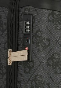 Valigia nera con motivo e zip decorati con doppio hardware dorato marchiato "GUESS?", e un lucchetto a combinazione nero integrato con logo TSA.