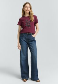 T-shirt bordeaux avec un motif floral noir, associé à un jean bleu taille haute à jambes larges. Texture du tissu naturel visible.