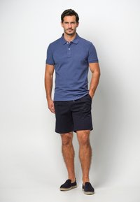 Mörkblå polo skjorta med korta ärmar, tvåknappars framknäppning, matchad med mörka shorts och mörka slipskor. Enkel, avslappnad outfit.