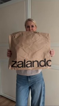 Rjava papirnata nakupovalna vreča z gubano teksturo, ki vsebuje velik črn logo "zalando". Oseba, ki drži vrečo, nosi sivo majico in modre kavbojke.
