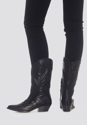 Botas de vaquero de cuero negro hasta la mitad de la pantorrilla con bordados de estrellas y llamas, combinadas con pantalones negros ajustados sobre un fondo neutro.