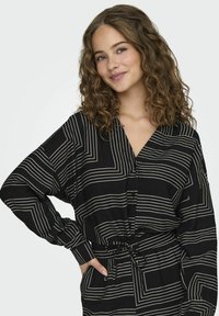 Zwarte shirt met witte geometrische lijnen, lange mouwen, V-hals, cinched taille, gemaakt van lichte stof met een gestructureerde afwerking.