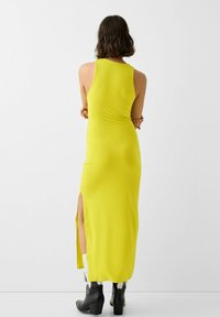 Žluté maxi šaty bez rukávů se bočním rozparkem; hladká textura; přiléhavý střih; nošeno s černými koženými botami na podpatku.