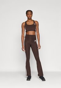 Καφέ κολάν με λευκές ρίγες στο πλάι και ασορτί αθλητικό σουτιέν. Λείος υφασματικός, εφαρμοστή σχεδίαση, και λογότυπο Adidas πάνω και κάτω.