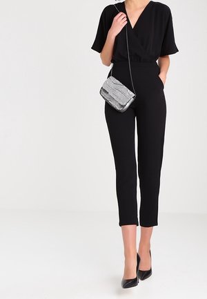 Femme portant une combinaison noire à manches courtes, col en V, pantalon court, talons noirs et un petit sac bandoulière argenté.