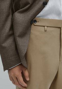 Blazer marrone con texture e due bottoni neri, abbinato a pantaloni beige con frontale piatto e tasche laterali, mani appoggiate sui fianchi.