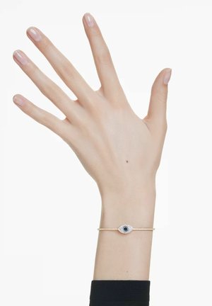 Linke Hand mit blasser Haut, die ein goldenes Armband trägt, das einen augenförmigen Anhänger mit dunklem Zentrum und umliegenden Kristallen zeigt.