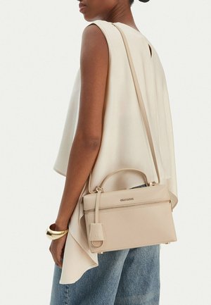 Donna con top crema senza maniche e jeans blu che porta una borsa beige Gino Rossi con tracolla e un braccialetto d'oro al polso.