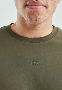 Butelkowozielony bawełniany t-shirt z okrągłym dekoltem. Na piersi subtelne, teksturowane logo QS. Gładka tkanina o dopasowanym kroju.