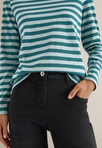 Person med hånden i lommen på sorte jeans iført en teal og lys blå stribet langærmet skjorte foran en ensfarvet væg.