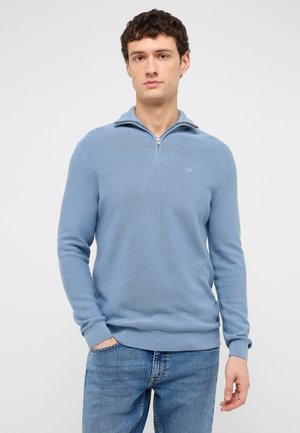 Junger Mann mit dunklem lockigem Haar, der einen hellblauen Pullover mit Viertelreißverschluss und blaue Jeans vor einem einfarbigen hellen Hintergrund trägt.