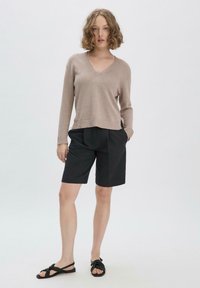 Beige Pullover mit V-Ausschnitt und geripptem Saum, kombiniert mit schwarzen maßgeschneiderten Shorts. Das Model trägt schwarze Sandalen mit Knoten-Detail. Einfaches, lässiges Outfit.