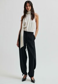 Czarne spodnie szerokie z gumką i mankietami na kostkach, zestawione z beżową bluzką bez rękawów z wysokim dekoltem i długim detalem w postaci szalika.