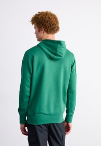 Sudadera con capucha verde de tela texturizada, de corte regular y con un bolsillo canguro. Presenta puños y dobladillo de canalé. Combinada con pantalones cargo oscuros.