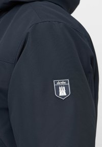 Giacca blu navy realizzata con un tessuto liscio, con un logo a scudo bianco e blu sulla manica sinistra che raffigura un'illustrazione di una torre.
