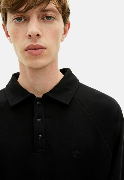 Jaunas vyras su trumpais rudais plaukais, vilkintis juodą marškinėlį su apykakle, turinčiu tris mygtukus ir subtilų siuvinėtą logotipą ant krūtinės.