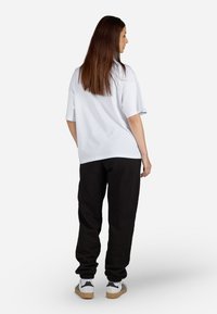 Tricou alb supradimensionat cu umeri lăsați, purtat cu pantaloni de trening negri cu manșete elastice. Modelul este întors cu fața la noi, purtând adidași.