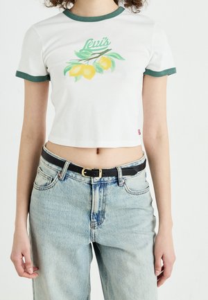 Biała krótka koszulka z zielonym obszyciem przy dekolcie, ozdobiona grafiką z cytryną i liśćmi. W połączeniu z jasnoniebieskimi dżinsami i czarnym paskiem.