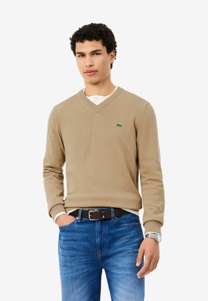 Joven con cabello rizado, vestido con suéter beige con cuello en V, jeans azules, cinturón negro y reloj de pulsera plateado, de pie frente a un fondo blanco.