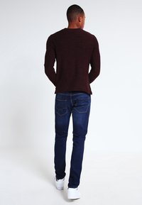 Mörkröd långärmad stickad tröja kombinerad med mörkblå raka jeans och vita sneakers, sedd bakifrån med minimal bakgrund.