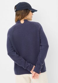 Navyblauwe trui met een gestructureerde brei, raglanmouwen en een klein sleutelgatdetail op de rug. Gedragen met een donkere baseballpet.