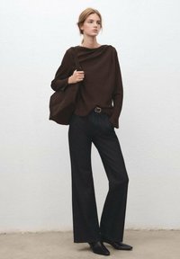 Hnedý dlhý top s dlhými rukávmi s voľným padnutím, kombinovaný s tmavými širokými džínsami. Doplnky zahŕňajú veľkú kabelku na rameno a čierne členkové čižmy so špicatou špičkou.