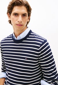 Πουλόβερ με ναυτικό μπλε και λευκές ρίγες, με στρογγυλή λαιμόκοψη, συνδυασμένο με πουκάμισο με ρίγες ανοιχτού γαλάζιου και γιακά από μέσα. Διαθέτει μικρή λεπτομέρεια με λογότυπο.