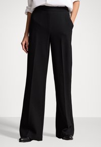 Osoba oblečená v černých kalhotách s širokými nohavicemi a černých botách, stojící s jednou rukou v kapse před jednobarevným pozadím.