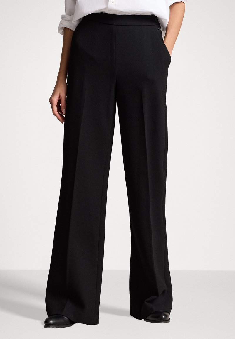 Osoba oblečená v černých kalhotách s širokými nohavicemi a černých botách, stojící s jednou rukou v kapse před jednobarevným pozadím.