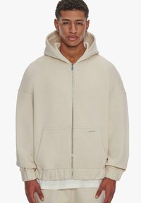 Beige huppari, jossa on vetoketju edessä, ylisuuri malli, ribbed hihansuut ja kengurutasku; pehmeä kangas, jossa on sileä pinta.