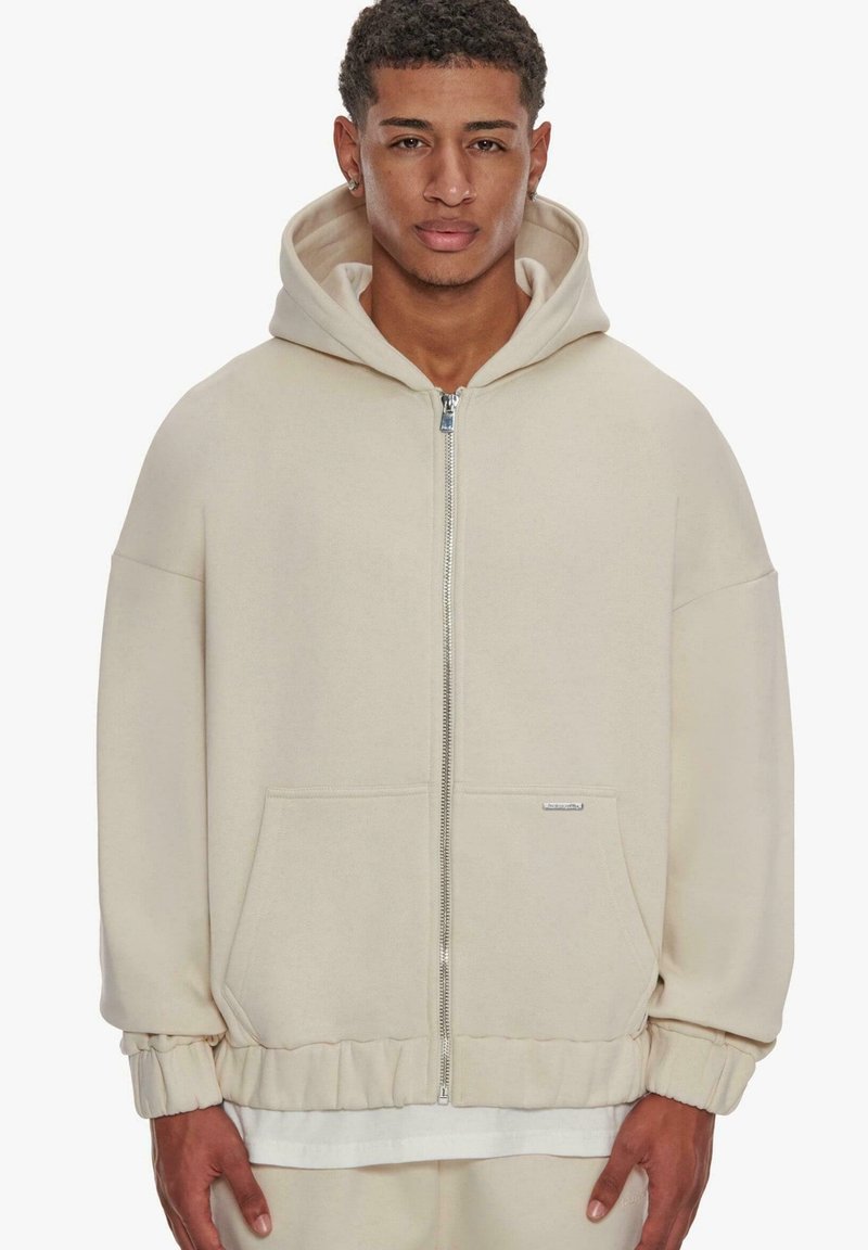 Beige huppari, jossa on vetoketju edessä, ylisuuri malli, ribbed hihansuut ja kengurutasku; pehmeä kangas, jossa on sileä pinta.