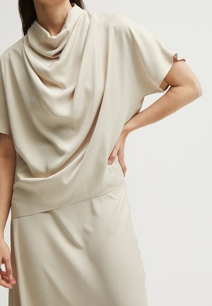 Kobieta w luźnej, kremowej bluzce z krótkim rękawem i drapowanym dekoltem oraz dopasowanej spódnicy, stojąca z jedną ręką na biodrze.