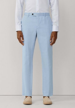 Hombre con pantalones azules claros a medida, alpargatas beige y camisa blanca de rayas de manga larga, de pie frente a un fondo liso.