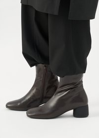 Botine din piele maro, cu un finisaj lucios, vârf rotund, fermoar pe lateral și toc gros negru. Purtați cu pantaloni negri largi.