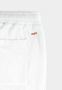 Vita ribbade joggers med elastisk midja, sidoficka och en liten orange broderad logotyp på baksidan.