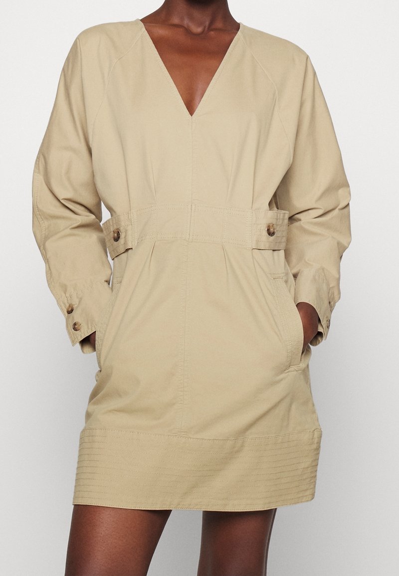 Robe mini beige à manches longues avec décolleté en V, pattes de taille à boutons, boutons aux poignets et poches latérales, portée avec les mains dans les poches.