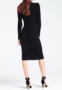 Femme aux longs cheveux roux portant une robe midi noire côtelée ajustée et des chaussures à talons hauts noires avec des brides à la cheville, debout devant un fond blanc.