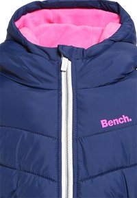 Giacca imbottita blu navy con fodera in pile rosa. Presenta una cerniera bianca e un logo visibile in rosa sul davanti. Texture trapuntata su tutta la superficie.