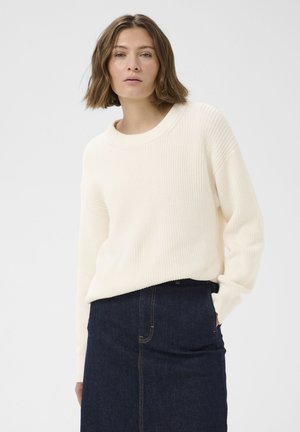 Vrouw draagt een witte gebreide trui en een donkerblauwe denim rok, staat met één hand in haar zak tegen een effen lichte achtergrond.