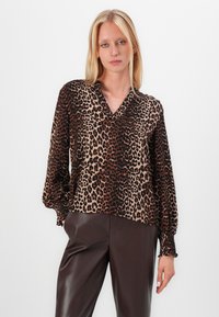 Blouse en imprimé léopard avec décolleté en V, manches longues à volants et poignets élastiques, fabriquée dans un tissu lisse et léger. Associée à un pantalon marron.
