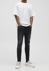Person som bär en vit oversized t-shirt, svarta slitna slim fit-jeans och vita sneakers med grå och gula detaljer, stående med händerna i fickorna.