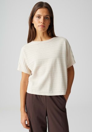 Femme aux longs cheveux bruns portant un haut texturé crème à manches courtes et un pantalon marron foncé, debout devant un fond clair uni.