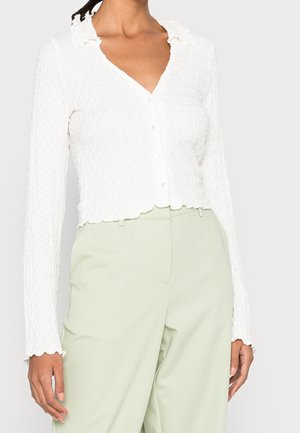 Personne portant un haut blanc texturé à manches longues boutonné avec des bords festonnés et un pantalon taille haute vert clair.