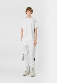 Camiseta de algodón blanca con escote rectangular, combinada con pantalones de chándal blancos que tienen detalles gráficos en negro y un corte ajustado. Botas con suela de goma.