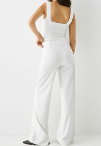 Donna che sistemando dei pantaloni a vita alta bianchi e larghi, indossando un top senza maniche bianco, in piedi contro uno sfondo luminoso e uniforme.