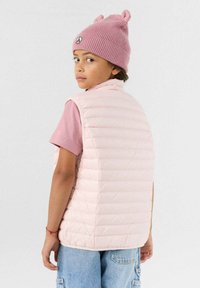 Lichtroze gewatteerde bodywarmer met een hoge kraag, gedragen over een roze T-shirt met korte mouwen. Het kind draagt ook een roze gebreide muts met oorflappen.