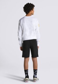 Camicia bianca a maniche lunghe, pantaloni corti neri, calzini neri con stampe grafiche e scarpe slip-on a scacchi, viste da dietro.